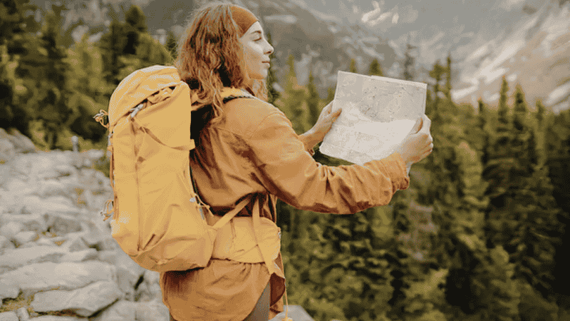 A beginner hiker standing at a trail junction, carefully checking a physical topographical map with a steep mountain trail and pine forest in the background.