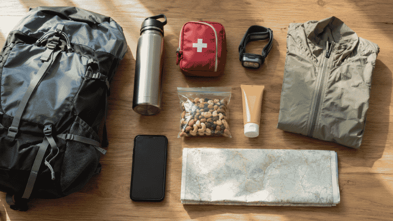 Flat lay of essential hiking gear for beginners including water bottle, headlamp, first aid kit, and trail snacks