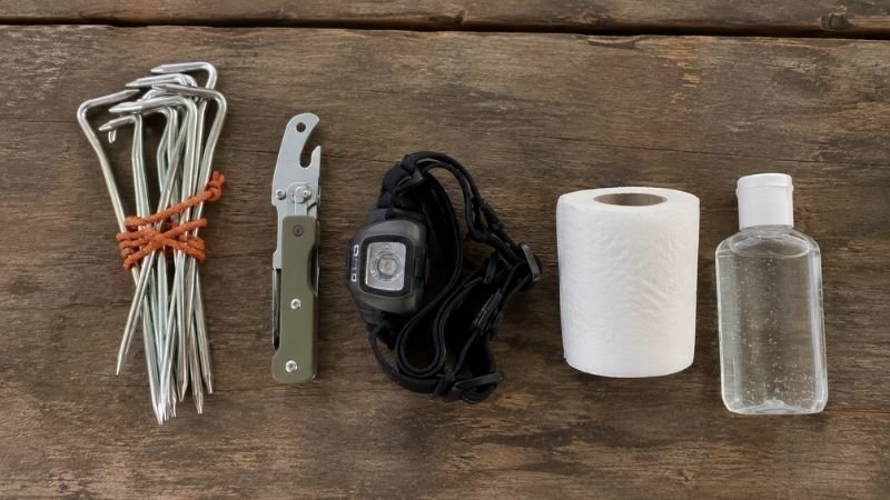 Flat lay of commonly forgotten camping items including tent stakes, can opener, headlamp, toilet paper, and hand sanitizer