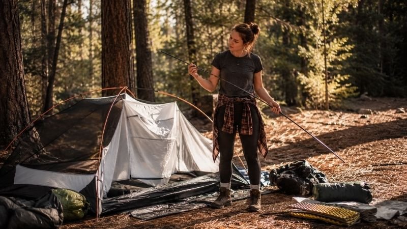 Young beginner camper standing beside a partially set up tent at a forest campsite holding a tent pole ready to learn how to set up a tent